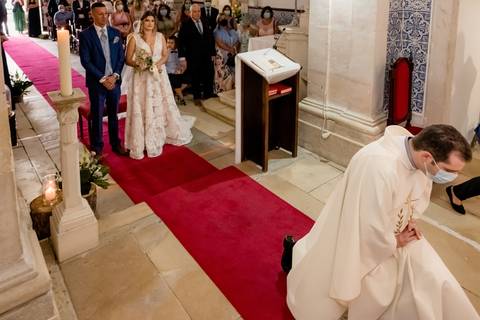 Uma noiva com umvestido de renda e um noivo com fato azul estão no altar em uma igreja, com um padre em vestes brancas ajoelhado em um tapete vermelho. Os convidados estão sentados e assistindo à cerimônia. Velas e arranjos florais decoram o corredor.'