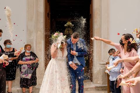 Um casal recém-casado sai da igreja enquanto os convidados os cobrem com arroz e pétalas. A noiva usa um vestido branco e segura um buquê, enquanto o noivo usa um fato azul.'