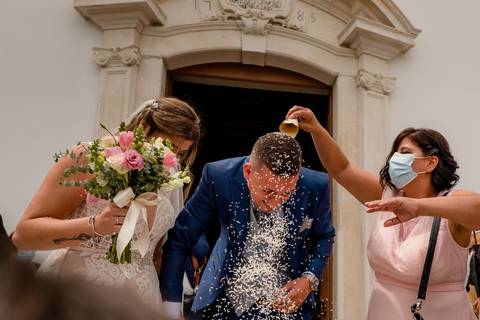 Um casal recém-casado sai da igreja enquanto os convidados os cobrem com arroz e pétalas. A noiva usa um vestido branco e segura um buquê, enquanto o noivo veste um fato azul.'