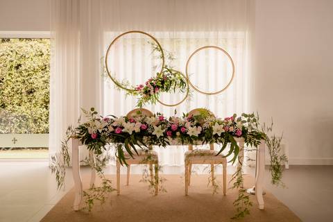Elegante recepção de casamento com a mesa dos  decorada com flores para dois em frente a cortinas transparentes. Acima, dois grandes anéis dourados adornados com flores rosas e brancas dão um toque romântico. A vegetação cai suavemente do arranjo da mesa.'