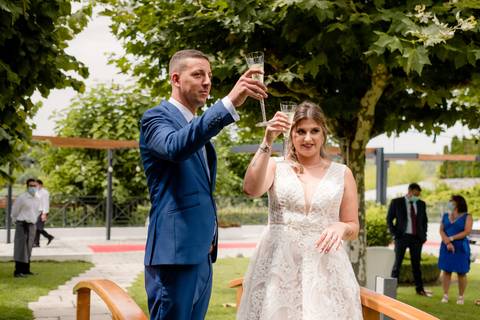Uma noiva em um vestido branco e um noivo com fato azul levantam suas taças para um brinde ao ar livre. Eles estão cercados por vegetação e um pequeno grupo de pessoas ao fundo.'