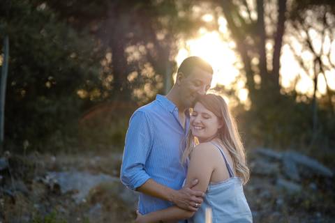 Um casal abraça-se ao ar livre ao pôr do sol, sorrindo de olhos fechados. Estão num cenário natural com árvores ao fundo.'