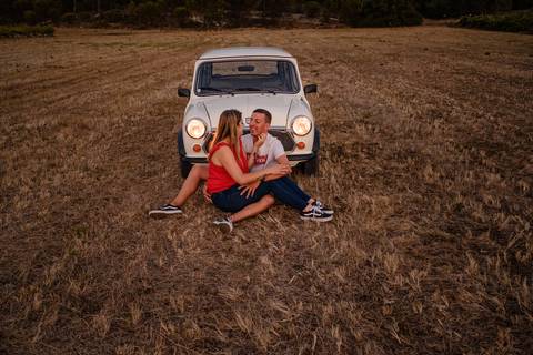 Um casal senta-se num campoem frente a um carro branco antigo num campo. Estão a sorrir um para o outro, com a mulher a usar um top vermelho e o homem uma camisa às riscas. A luz do entardecer projecta um brilho quente sobre a cena.'