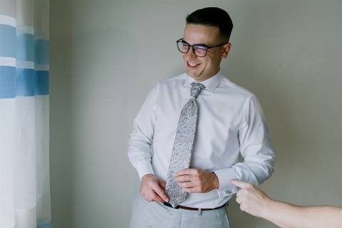 m momento descontraído do noivo enquanto ajusta a gravata, exibindo um sorriso genuíno. A luz suave e o ambiente simples tornam a fotografia autêntica e natural, capturando a leveza e alegria do dia.'