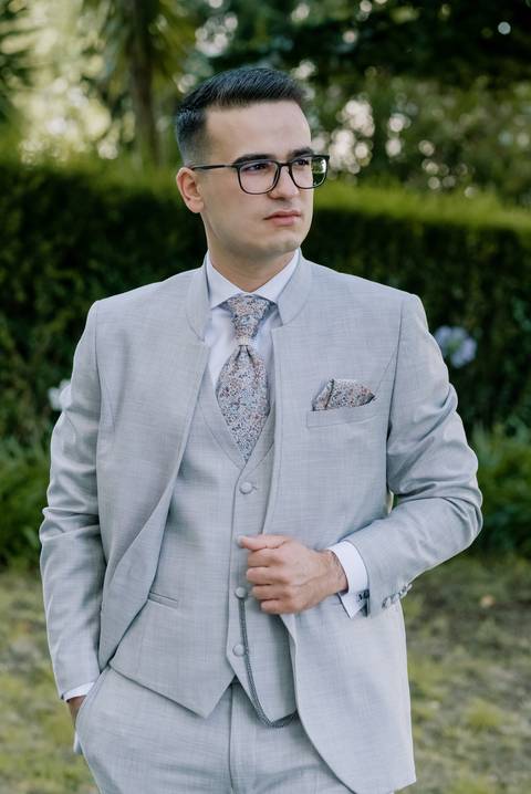Noivo jovem e elegante, vestindo um fato cinzento claro, com cabelo penteado e óculos. Ele usa uma gravata e um lenço no bolso com um padrão delicado, ajustando o casaco com uma das mãos. O cenário de fundo é verde e natural'
