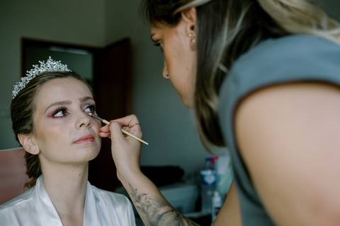 Um momento dos preparativos da noiva, capturado enquanto a maquilhagem final é cuidadosamente aplicada. A noiva, usando um robe de cetim branco e uma tiara elegante. Este retrato destaca a atenção aos detalhes.'