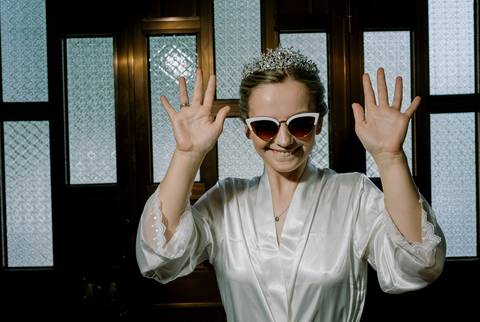 Um momento descontraído e cheio de personalidade da noiva. Vestindo um robe de cetim branco com detalhes em renda, a noiva exibe um sorriso contagiante, complementado por óculos de sol.'