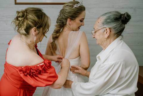 Momento emocionante do casamento: a noiva, com um vestido elegante e tiara, é ajudada pela mãe e pela avó enquanto ajustam os últimos detalhes do vestido. Cena cheia de carinho e tradição, ideal para inspirar noivas e destacar momentos familiares únicos.'