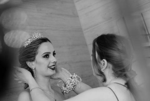 Nesta fotografia a preto e branco, a noiva sorri enquanto a sua irmã a ajuda. O momento capta a emoção e a elegância dos preparativos antes da cerimónia, destacando o brilho da noiva e a cumplicidade entre ambas.'