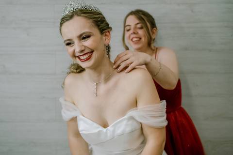 Na imagem, a noiva sorri radiante enquanto a irmã, vestida de vermelho, ajusta o colar delicado que complementa o vestido branco elegante. Um momento de cumplicidade e felicidade, perfeito para destacar os preparativos do casamento e a relação entre ambas'
