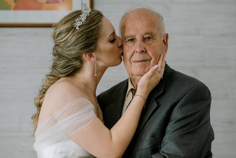 Noiva beija carinhosamente o avô durante a sua preparação. Com um vestido elegante e tiara, ela demonstra amor e ligação familiar. O avô, de fato cinzento, transmite serenidade. Momento especial que celebra laços entre gerações em um dia inesquecível.'