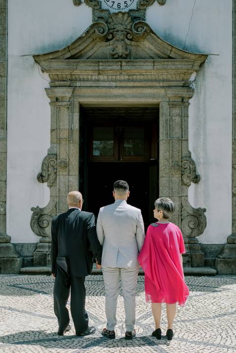 Noivo de fato cinzento caminha de mãos dadas com os pais, simbolizando amor e cumplicidade familiar. O pai usa fato preto elegante, e a mãe destaca-se com um vestido rosa vibrante, conferindo um toque de cor e sofisticação à ocasião especial do casamento.'