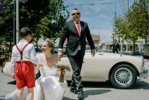 Noiva elegante, usando vestido branco clássico, cumprimenta carinhosamente uma criança. Ao fundo, o pai da noivacom fato preto e gravata vermelha complementa a cena, destacando um carro clássico.'
