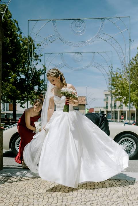 Noiva radiante, com vestido branco volumoso e véu longo, segura um delicado bouquet de flores brancas enquanto ajusta o vestido. Ao lado, a dama em vestido vermelho ajuda com os detalhes. Ao fundo, um carro clássico.'