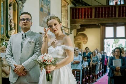 Noiva emocionada limpa uma lágrima durante a cerimónia de casamento, segurando um delicado ramo com flores brancas e vermelhas. Ao lado, o noivo, com fato cinzento claro e gravata elegante. Ao fundo, os convidados acompanham o momento.'