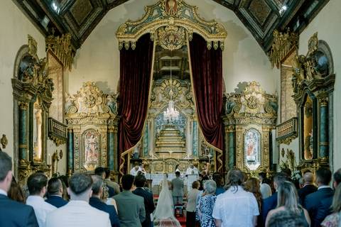 Interior de uma igreja histórica com altares dourados, cortinas de veludo vermelho e um lustre central. No altar, dois padres celebram a cerimónia, com a noiva e o noivo de frente para eles. Os bancos estão ocupados por convidados.'