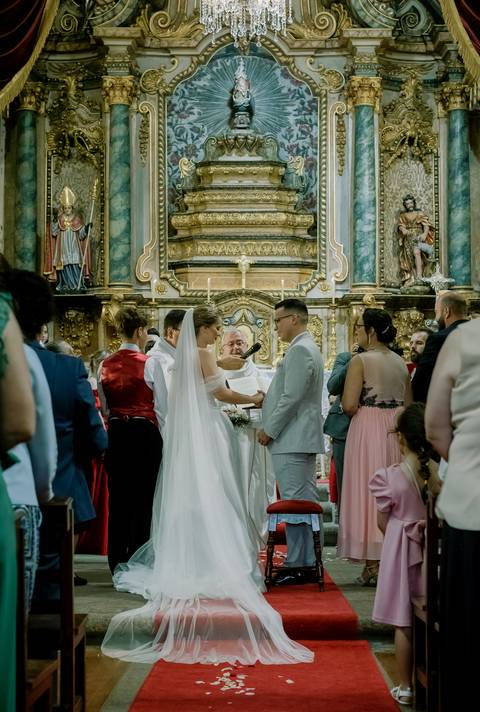 Casamento numa igreja histórica, com noivos trocando votos no altar ricamente decorado com dourados e imagens sacras. A noiva, de véu e vestido branco, e o noivo, de fato claro, recebem a bênção do sacerdote enquanto convidados assistem emocionados.'