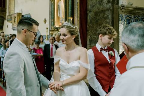 Casamento numa igreja histórica com destaque para os noivos. O noivo, com um fato cinzento, segura a mão da noiva, vestida com um clássico vestido branco, véu e uma tiara.'