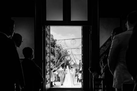 Fotografia de casamento a preto e branco, captada à saída de uma igreja em Portugal, com os noivos de mãos dadas. O contraste entre o interior sombreado e o exterior iluminado com arcos festivos tradicionais realça a emoção do momento. '