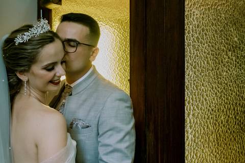Casal de noivos num momento íntimo e romântico capturado durante o casamento. A noiva, sorri enquanto recebe um beijo carinhoso do noivo. O fundo é composto por uma porta de madeira e vidro texturizado, criando um contraste acolhedor e refinado.'