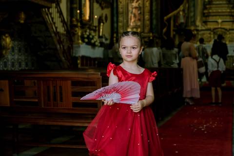 Menina das alianças vestida com um elegante vestido vermelho com detalhes brilhantes, segurando um leque decorativo, num ambiente de igreja. O fundo revela o altar decorado e convidados do casamento, destacando a atmosfera da cerimónia.'