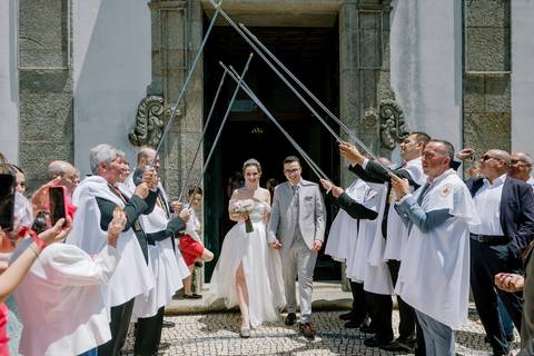 Saída dos noivos da igreja em Portugal, celebrada com uma guarda de honra. A noiva, com bouquet, e o noivo, sorridentes, são recebidos por familiares e amigos. Palavras-chave: casamento em Portugal, saída da igreja'