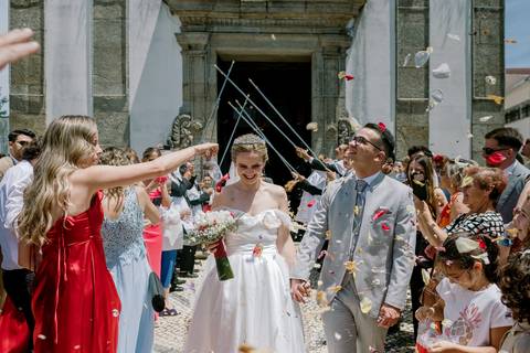 Momento especial de casamento em Portugal com os noivos sorridentes à saída da igreja, enquanto são recebidos com pétalas de flores e aplausos. Destaque para a felicidade dos noivos e a celebração ao ar livre. casamento em Portugal, saída da igreja'