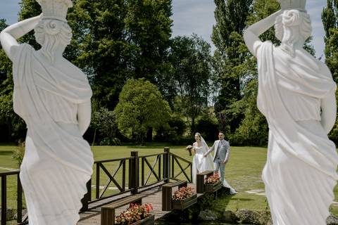 Noivos atravessam uma ponte de madeira cercada por estátuas clássicas e flores, em cenário de jardim ao ar livre ideal para casamentos. Espaço rodeado por natureza, perfeito para cerimónias únicas e memoráveis. casamentos  ar livre, quinta dos Jasmins '