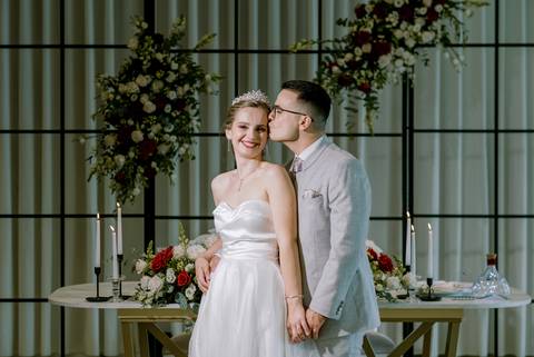 Momento romântico dos noivos durante o casamento, com decoração elegante de flores brancas e vermelhas, luz de velas e ambiente requintado. A noiva sorridente é beijada pelo noivo, transmitindo amor e felicidade.
casamento romântico, fotografia  Casamento'