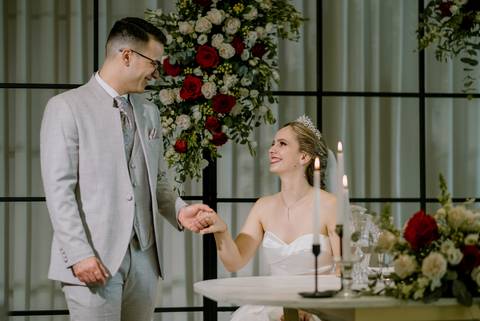 Momento especial entre os noivos no casamento, com decoração sofisticada de flores vermelhas e brancas e velas ao centro. A noiva, sorridente, segura a mão do noivo, transmitindo amor e cumplicidade, xcasamento elegante, fotografia de Casamento  '