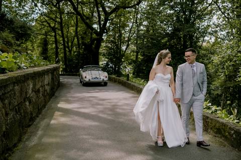 Noivos apaixonados caminham de mãos dadas numa estrada ladeada por natureza, com um carro clássico ao fundo. Momento de casamento único, repleto de elegância e romantismo. casamento, noivos, natureza, carro clássico, amor, sessão fotográfica.'