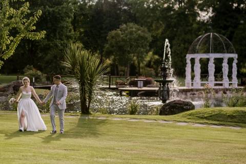 Noivos caminham de mãos dadas num cenário de sonho com jardim, fonte e lago ao fundo. Ambiente perfeito para fotografias de casamento, combinando elegância e natureza. casamento, noivos, jardim, fonte, lago, romantismo, fotografia de casamento.'