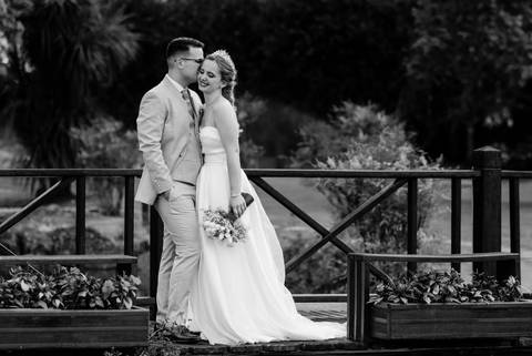 Noivos apaixonados em retrato preto e branco sobre uma ponte de madeira, transmitindo romantismo e elegância. Detalhes do vestido de noiva e do ramo de flores destacam-se. casamento, noivos, fotografia artística, ponte, romantismo, preto e branco.'