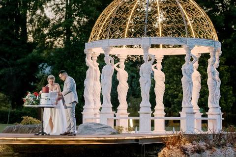 Noivos a cortarem o bolo de casamento sob uma elegante pérgola iluminada, rodeada por esculturas clássicas. Cenário requintado ideal para cerimónias ao ar livre. casamento, bolo dos noivos, pérgola, decoração de casamento, cerimónia ao ar livre.
'