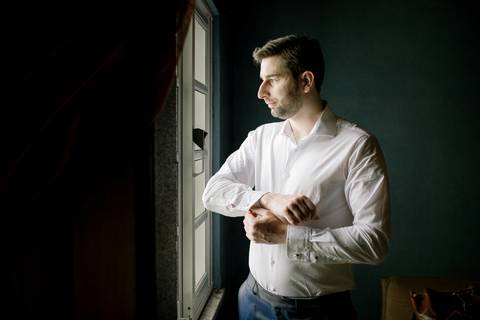 
Noivo ajusta os botões da camisa enquanto olha pela janela, num momento íntimo de preparação para o casamento. A fotografia realça elegância, luz natural e emoções genuínas. noivo, preparação, casamento, momento íntimo, fotografia de casamento'