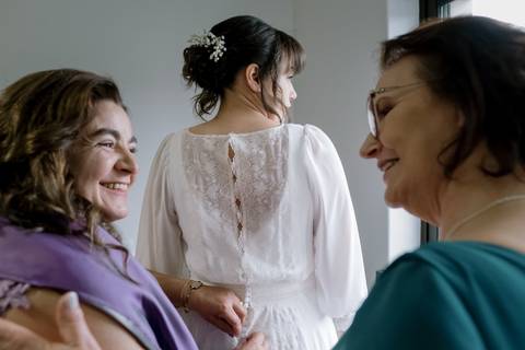 Noiva em preparação, com detalhe no vestido de renda e botões delicados, rodeada por familiares sorridentes. Momento íntimo e emotivo antes do casamento. noiva, preparação, vestido de renda, casamento, momentos especiais, tradição, Portugal..'