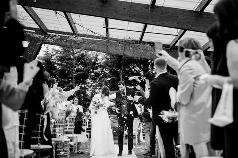 
Fotografia a preto e branco de um casal de noivos a caminhar pelo corredor após a cerimónia, rodeados por convidados a lançar pétalas e aplaudir. Momento cheio de alegria e emoção. casamento em Portugal, saída dos noivos, celebração ao ar livre.'