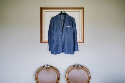 Casaco azul de fato de noivo pendurado num cabide, com detalhe de lenço no bolso, enquadrado por uma moldura elegante. Preparativos do noivo capturados com estilo e simplicidade. fato de noivo, casamento, preparativos, elegância masculina. '