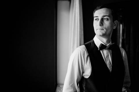 Retrato elegante do noivo a preto e branco, captado num momento de introspeção junto a uma janela, iluminado por luz natural. O noivo veste um colete e laço num estilo clássico. Fotografia, 
Retrato de casamento, otógrafo profissional em Portugal'