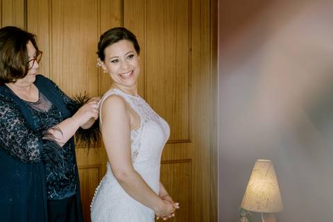 Noiva sorridente é ajudada pela mãe a abotoar o vestido de renda no dia do casamento. Um momento íntimo e especial, capturado com detalhes emotivos e luz suave no quarto. noiva,  casamento, vestido de noiva, fotografia de casamento, preparação da noiva.'