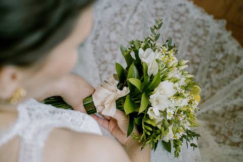 Detalhe do bouquet da noiva com flores frescas em tons de branco e amarelo, complementado por folhagens verdes e um laço delicado. A composição destaca a elegância e a beleza natural do momento do casamento.  bouquet de noiva, casamento'
