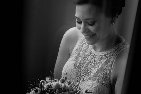 Fotografia de noiva sorridente em preto e branco, com vestido de renda delicado e bouquet floral. A imagem captura a emoção e elegância da noiva no dia do casamento. noiva, vestido de renda, bouquet de noiva, casamento, fotografia de casamento.'