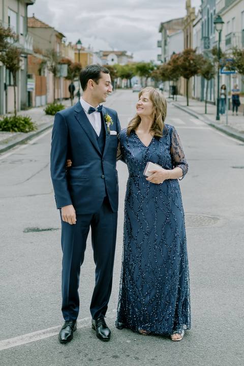 Fotografia de noivo sorridente ao lado da mãe no dia de casamento. Ambos elegantes, o noivo com fato azul  e a mãe com vestido azul com brilho. Ambiente descontraído em rua pitoresca. noivo, mãe do noivo, casamento, fato azul.'