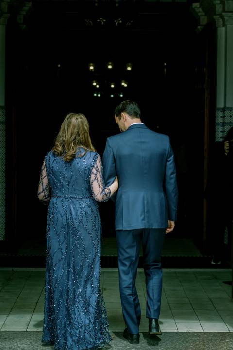 Mãe e noivo, de costas, a caminhar juntos para a cerimónia, num momento cheio de significado e emoção. A mãe usa um vestido azul elegante com detalhes bordados. noivo, casamento, cerimónia, fotografia, momento especial.'