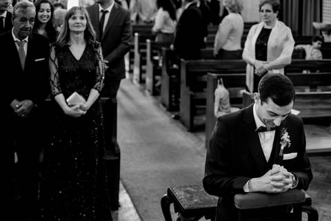 Noivo ajoelhado em momento de oração na igreja, com a mãe em destaque na fila da frente, usando um vestido elegante. Fotografia a preto e branco que capta a emoção e solenidade da cerimónia. casamento, noivo, mãe do noivo, igreja, fotografia emotiva.'