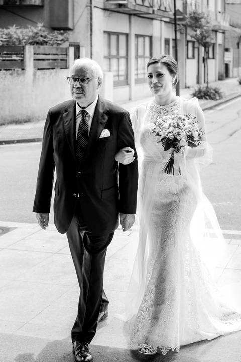 Noiva acompanhada pelo pai, prestes a entrar na igreja, captada em fotografia a preto e branco. O vestido rendado e o bouquet destacam a elegância e emoção do momento. casamento, noiva, pai da noiva, entrada na igreja, fotografia de casamento.'