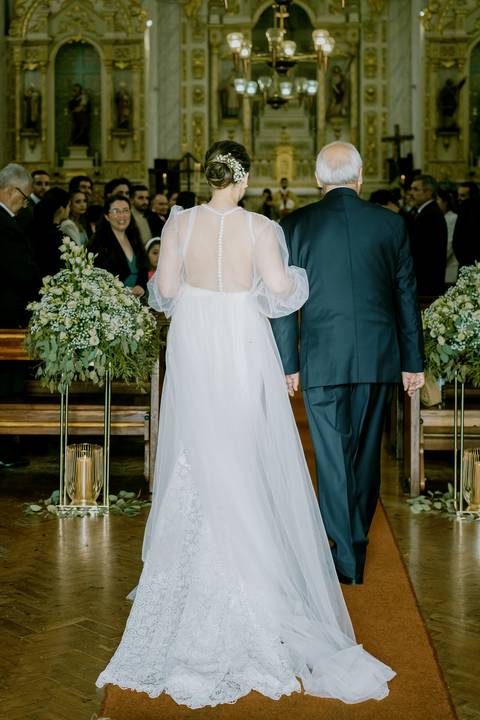 Noiva acompanhada pelo pai caminha pelo corredor da igreja decorada com flores elegantes, rumo ao altar. A fotografia destaca o vestido de noiva rendado e a emoção do momento. casamento, entrada na igreja, noiva, fotografia de casamento.'
