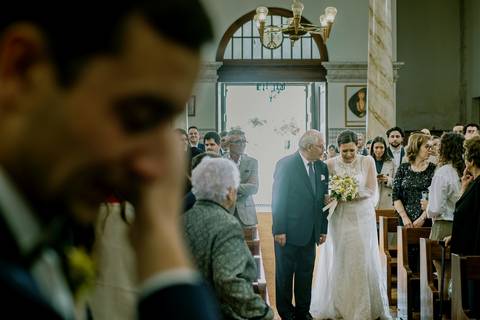 Noiva entra na igreja com o pai enquanto o noivo, emocionado, reage ao momento. A fotografia captura a emoção e o amor no casamento, rodeados por familiares e amigos. casamento, noiva, noivo emocionado, entrada na igreja, fotografia de casamento.'