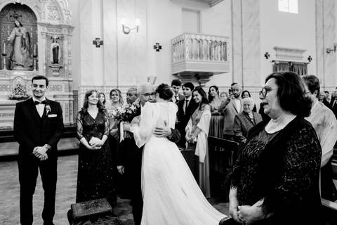 Fotografia de casamento em preto e branco na igreja, onde a noiva abraça o pai com emoção enquanto o noivo observa sorridente. Detalhes do altar e convidados presentes enriquecem o momento. casamento, noiva, noivo, igreja, fotografia emocional.'