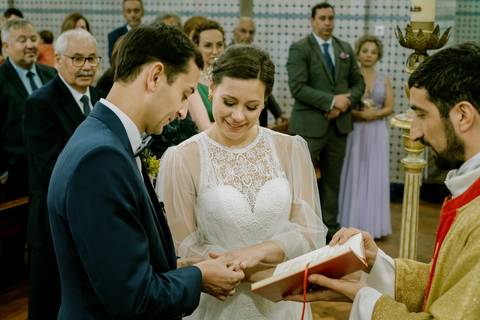 Troca de alianças durante a cerimónia de casamento: a noiva e o noivo partilham um momento de amor e emoção enquanto o padre celebra o ritual. Fundo com convidados atentos. casamento, troca de alianças, cerimónia, noiva, noivo, fotografia profissional.'